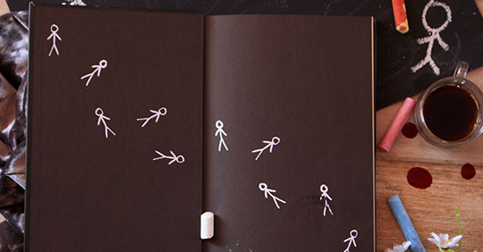 Livro o Homem de Giz aberto mostrando a primeira página com vários homens de giz desenhados, o livro está sobre uma mesa com vários pedaços de giz e um copo com café.