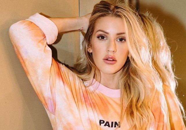 Ellie Goulding com blusa laranja e mão na cabeça