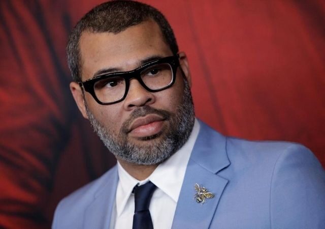 Jordan Peele sério com óculos de grau preto e terno azul, blusa branca e gravata preta
