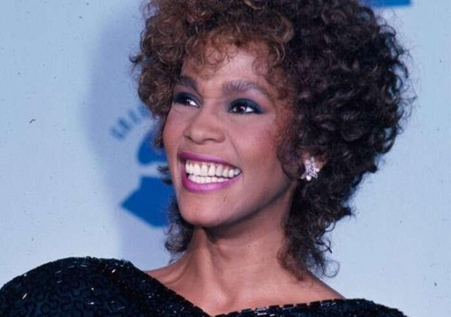 Whitney Houston sorri e usa roupa preta, tem cabelos cacheados e curtos