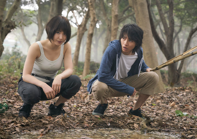 Imagem da série Alice in Borderland, com duas pessoas em uma floresta com muitas folhas secas no chão. 