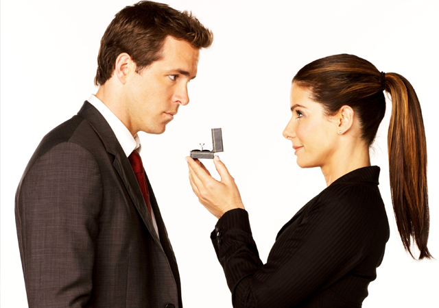 Recorte do pôster do filme A Proposta. Mulher segurando uma caixinha com anel de noivado para um homem que a encara.